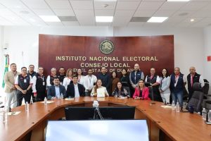 más de 10 personas de pie frente a una mesa ovalada color madera