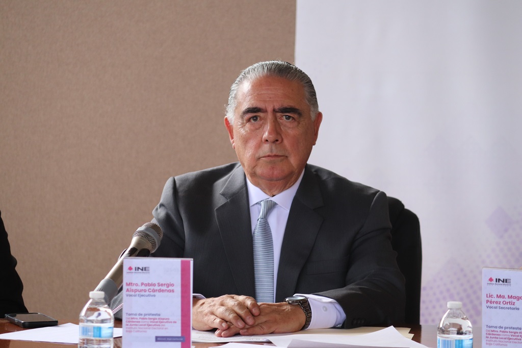 Hombre con pelo cano vestido de traje sentado detrás de un micrófono