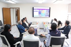 Nueve personas sentadas al rededor de una mesa larga frente a una pantalla