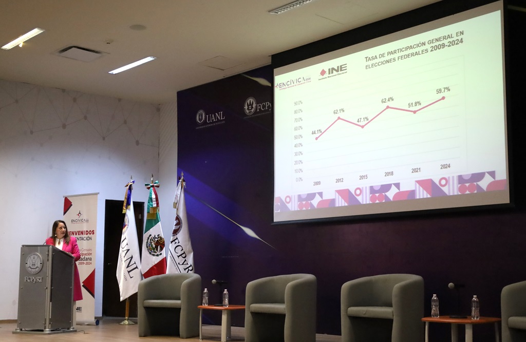 mujer de pie con vestido rosa hablando a través de un micrófono junto a una pantalla