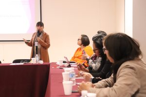 cuatro personas sentadas y una personas de pie detrás de una mesa hablando a través de un micrófono