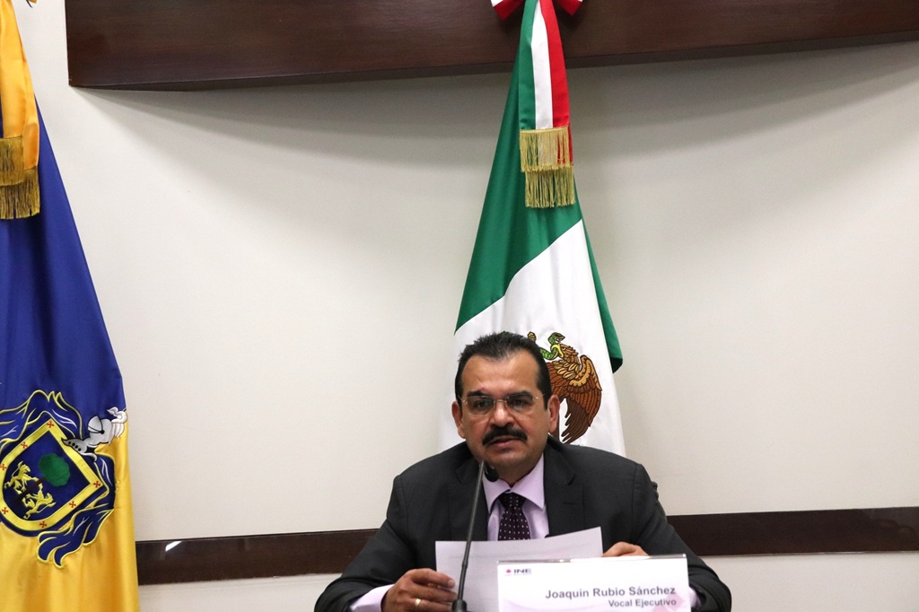 hombre sentado, bigote con lentes, leyendo frente a un micrófono