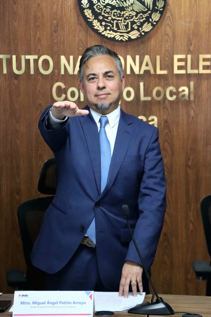 hombre cabello cano, traje azul, de pie con el brazo derecho extendido al frente