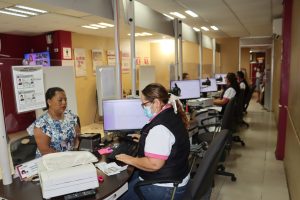 Una mujer con cabello recogido y cubrebocas sentada en una mesa frente a una computadora atendiendo a otra mujer vestida de blanco y azul.