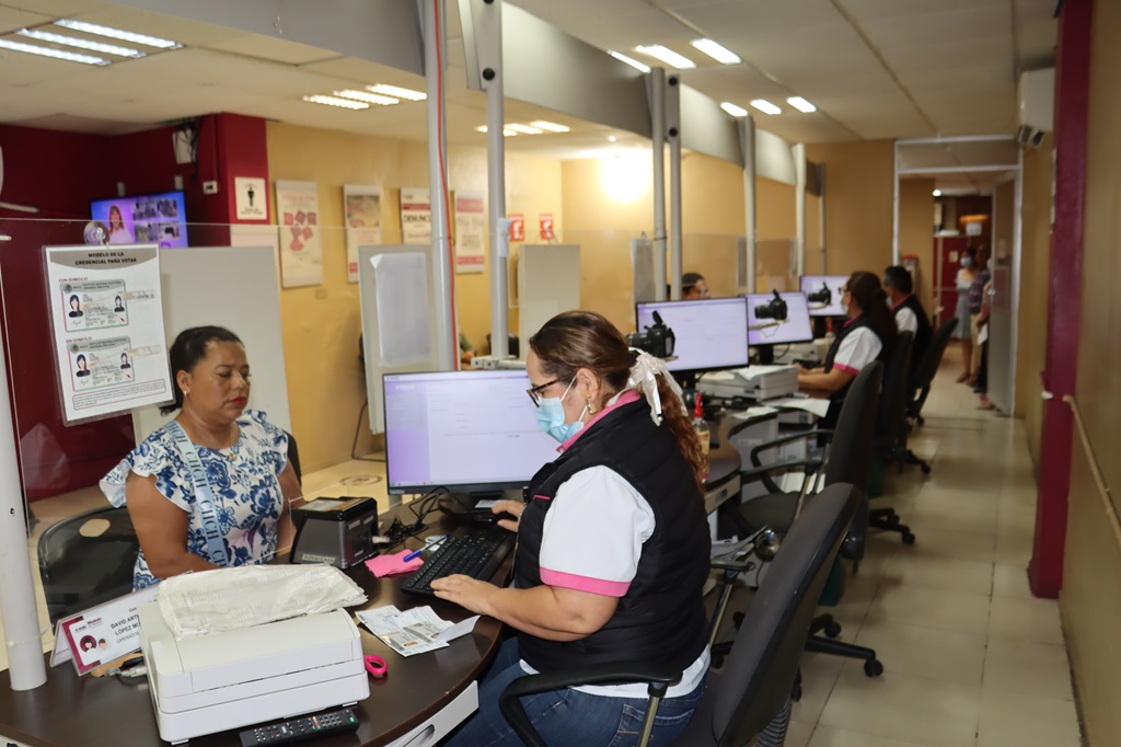 Una mujer con cabello recogido y cubrebocas sentada en una mesa frente a una computadora atendiendo a otra mujer vestida de blanco y azul.