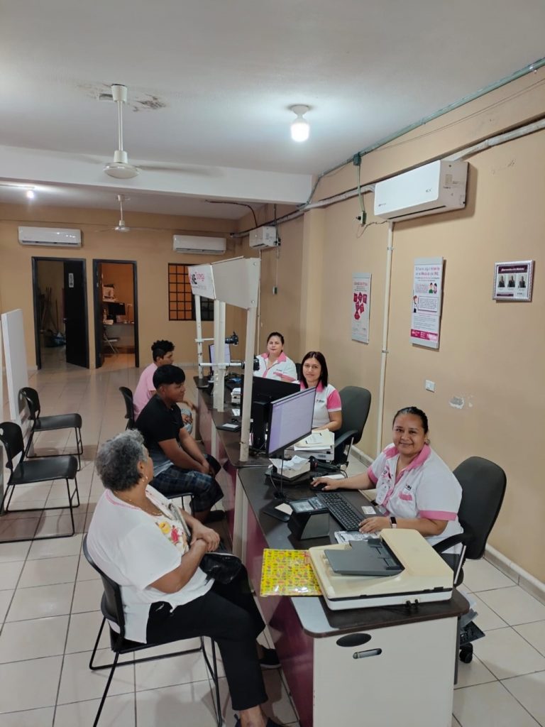 Tres mujeres sonrientes, sentadas en una mesa frente a computadoras atendiendo a la ciudadanía