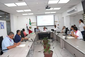 Nueve personas reunidas, sentadas detrás de una mesa larga, hablando detrás de micrófonos.
