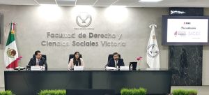 tres personas sentadas frente a una mesa y a los costados dos banderas