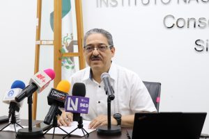 un hombre pelo cano, lentes, camisa blanca, sentado frente a siete micrófonos