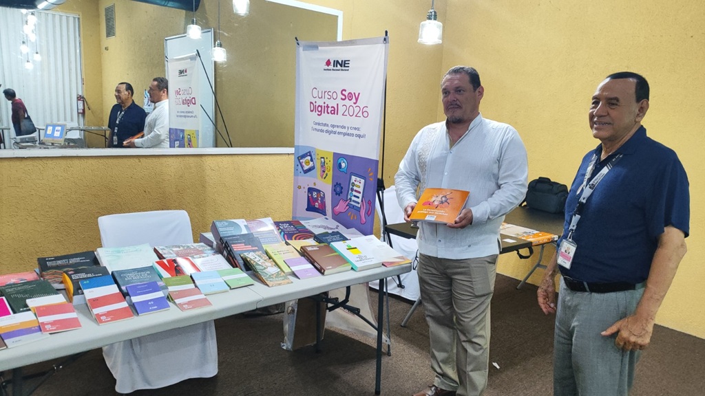 Dos hombres de pie a un costado de una mesa con muchos libros de diferentes colores