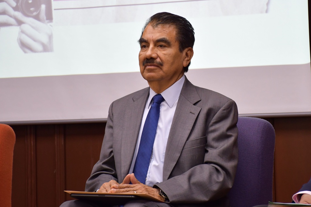 Un hombre sentado con saco gris, camisa blanca y corbata azul
