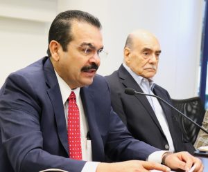 dos hombre mayores sentados, uno de ello habla por un micrófono