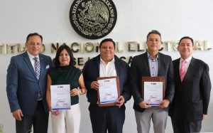 una mujer y cuatro hombres de pie, sostienen un diploma.