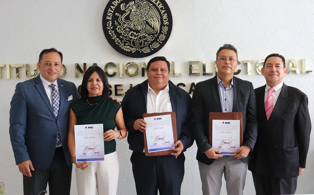 una mujer y cuatro hombres de pie, sostienen un diploma.