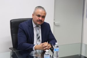 Hombre con cabello cano y bigote, vestido de traje negro y corbata, sentado detrás de una mesa