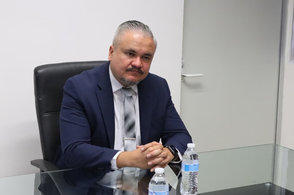 Hombre con cabello cano y bigote, vestido de traje negro y corbata, sentado detrás de una mesa