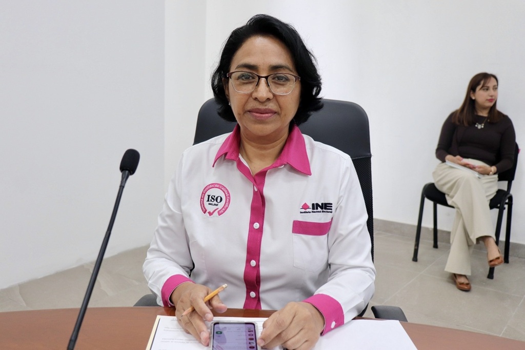 una mujer sentada, cabello corto viste camisa blanca con rosa y trae lentes