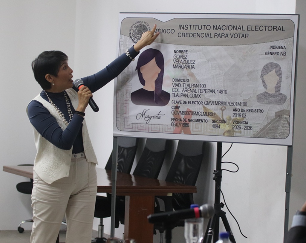 persona de pie cabello corto señalando una imagen gigante de la credencial para votar del INE