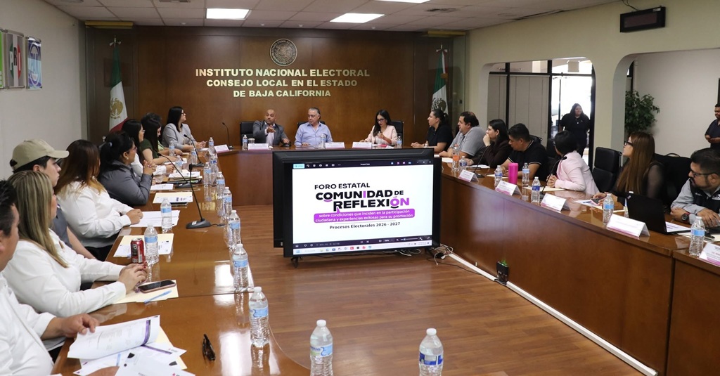personas sentadas en un salón con mesa ovalada, en medio una pantalla de Televisión