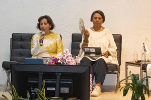 Dos mujeres sentadas, una de ellas habla por micrófono