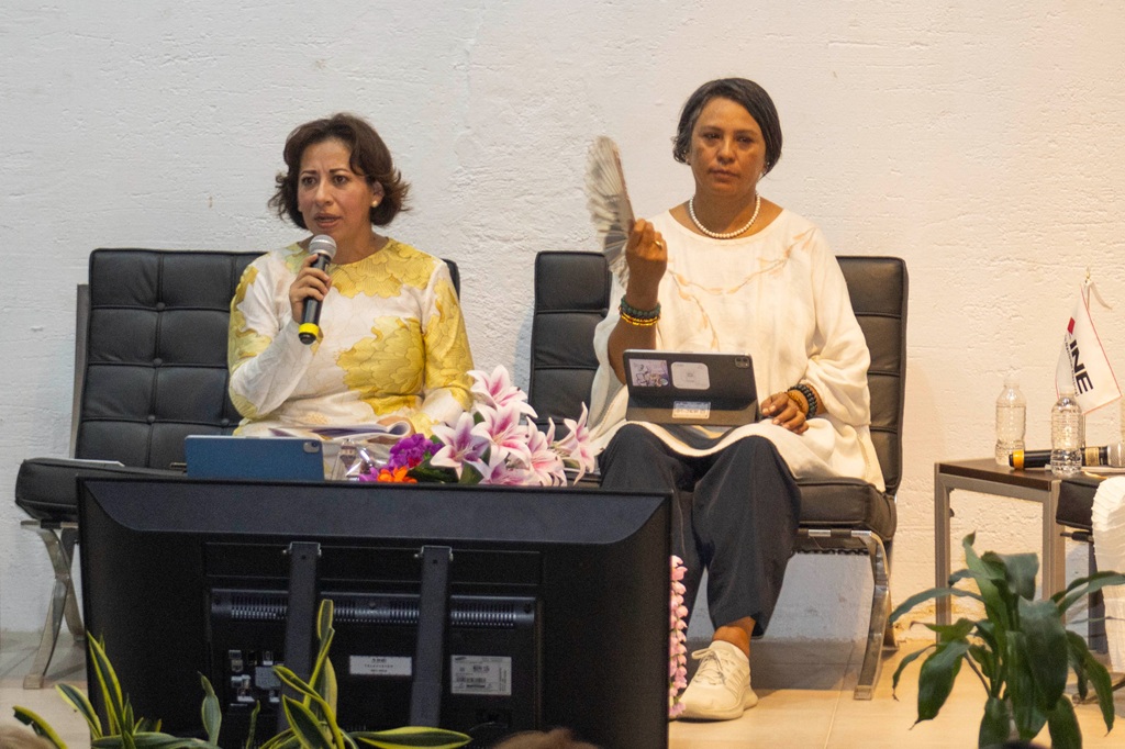 Dos mujeres sentadas, una de ellas habla por micrófono