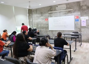 personas sentadas en un salón de clases frente a un pizarrón blanco