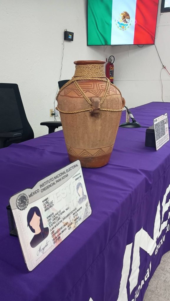 Una bandera de México, una mesa con mantel morado, un jarrón de barro y una credencial para votar