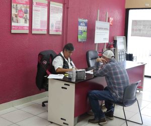 una adulta mayor sentada revisando papeles, frente a ella un hombre con gorrra sentado