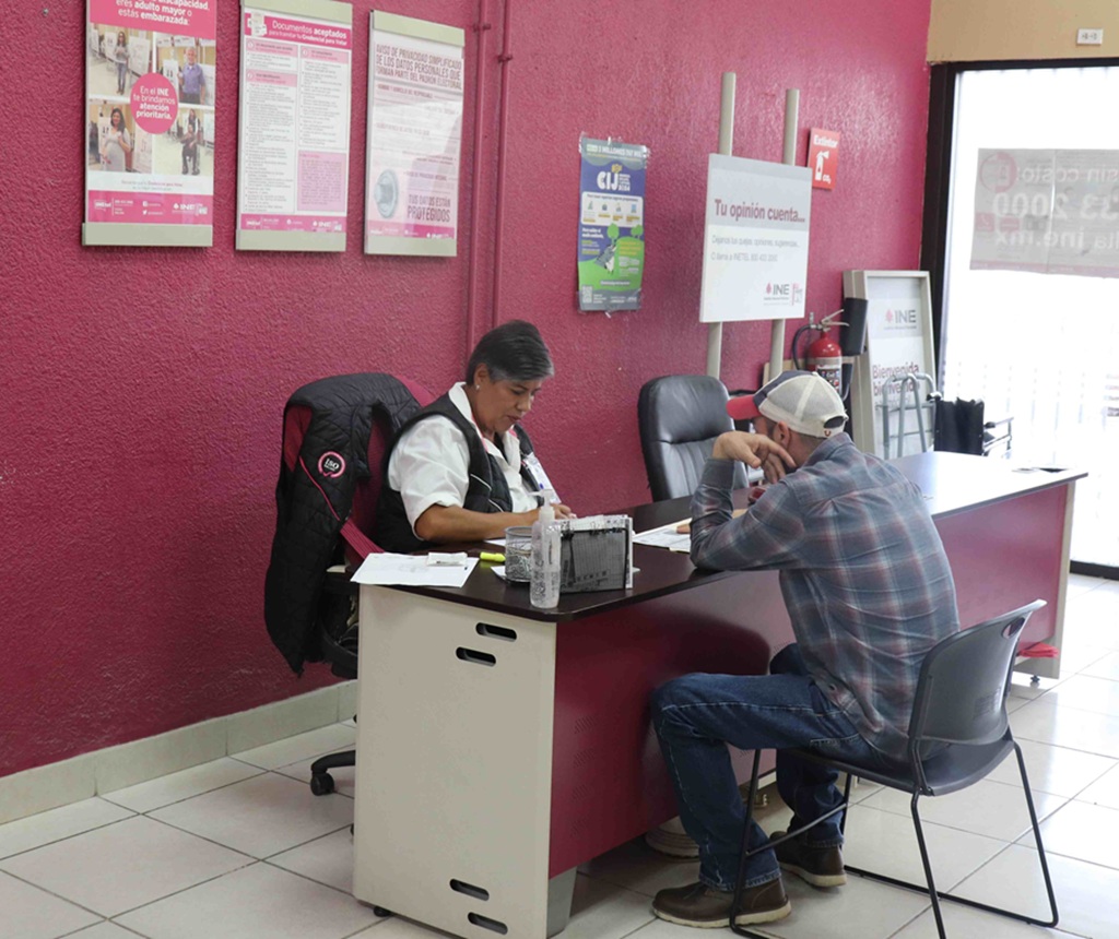 una adulta mayor sentada revisando papeles, frente a ella un hombre con gorrra sentado