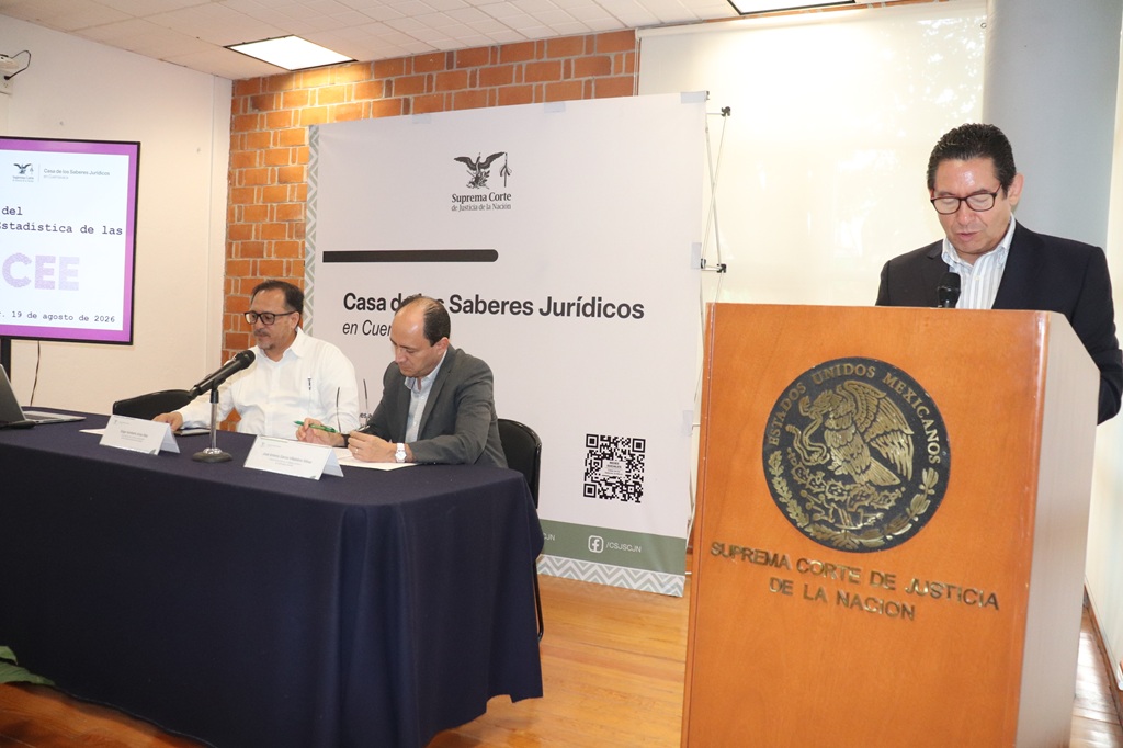 tres hombres, uno de pie frente a un micrófono y dos sentados a su costado