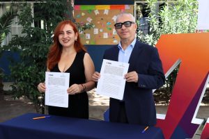 una mujer cabello rojizo largo y vestido negro de pie, a un lado un hombre con lentes, ambos sostienen un documento