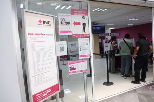 entrada de una oficina, tiene perta de cristal, y letreros color blanco y rosa con letras negras que dicen INE