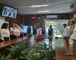varias personas de pie en un salón frente a cinco estudiantes uniformadas sosteniendo una bandera nacional