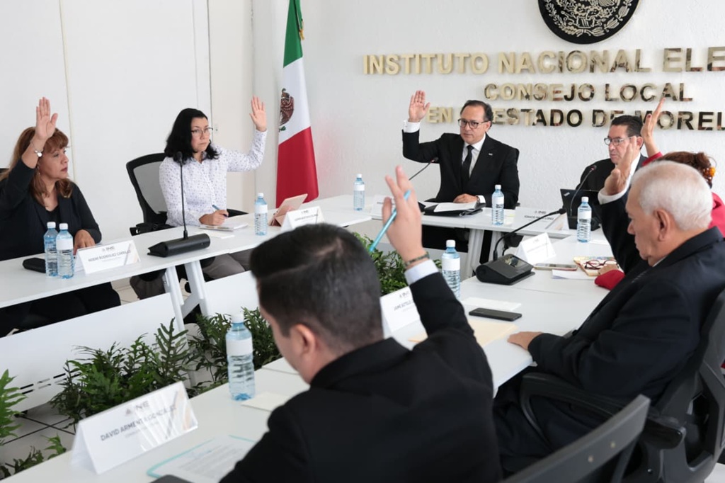 varias personas en un salón blanco, sentadas votando a mano alzada