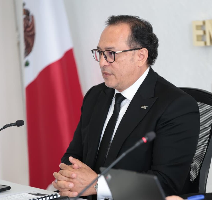hombre de traje negro y lentes, sentado frente a un micrófono