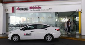 un carro blanco con un letrero del INE estacionado afuera de un módulo