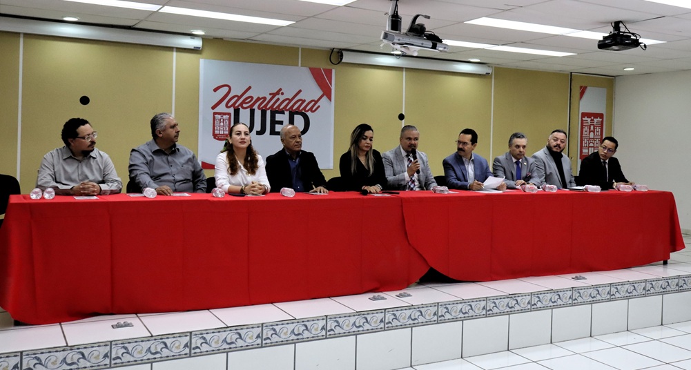 Dos mujeres y ocho hombres sentados frente a una mesa con mantel rojo y micrófonos