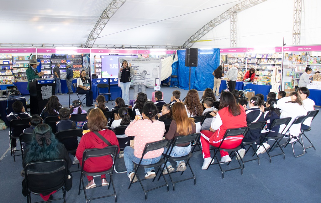 personas menores sentadas, alrededor muchos libros, escuchando a un tres personas en un escenario
