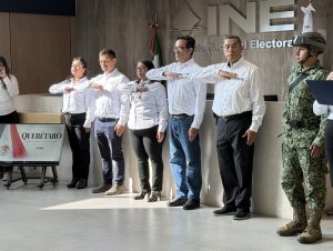 dos mujeres y tres hombres de pie, vestidos de pantalón negro y camisa blanca con el brazo derecho en el pecho viendo la bandera nacional