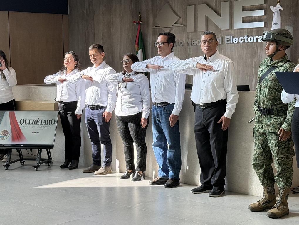 dos mujeres y tres hombres de pie, vestidos de pantalón negro y camisa blanca con el brazo derecho en el pecho viendo la bandera nacional