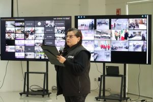 Mujer vestida de negro y de pie frente a varias pantallas dando un mensaje