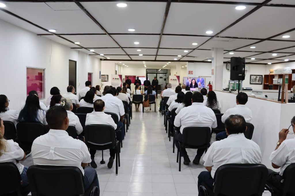 varias personas con camisa blanca sentados viendo de frente a una oradora en pantalla