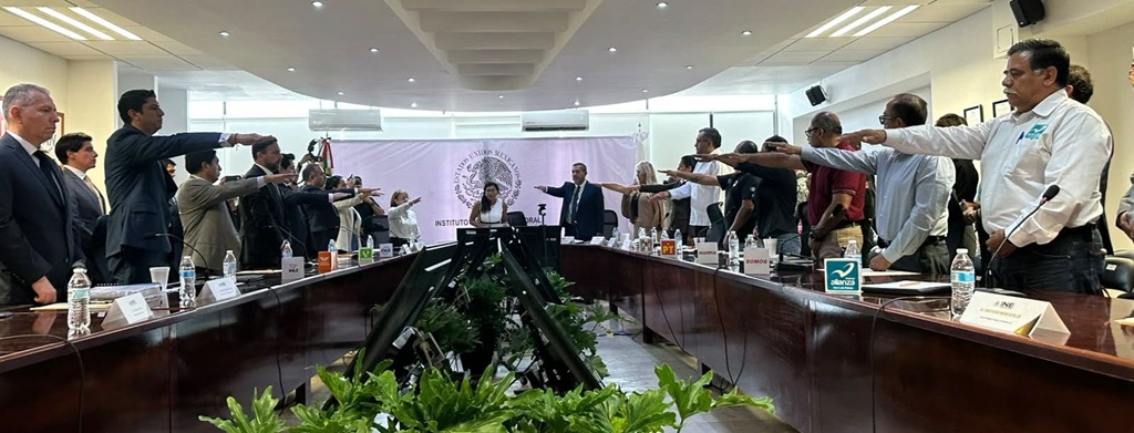 varias personas de pie alrededor de una mesa rectangular con el brazo entendido al frente