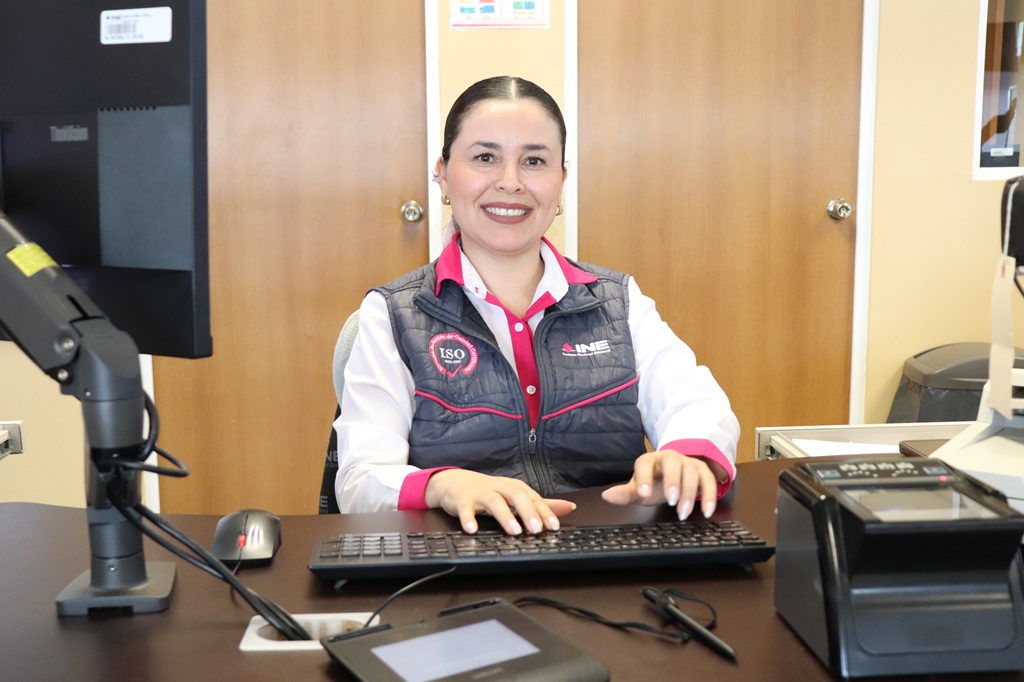 Mujer cabello recogido y con uniforme gris con rosa sentada viendo a la cámara