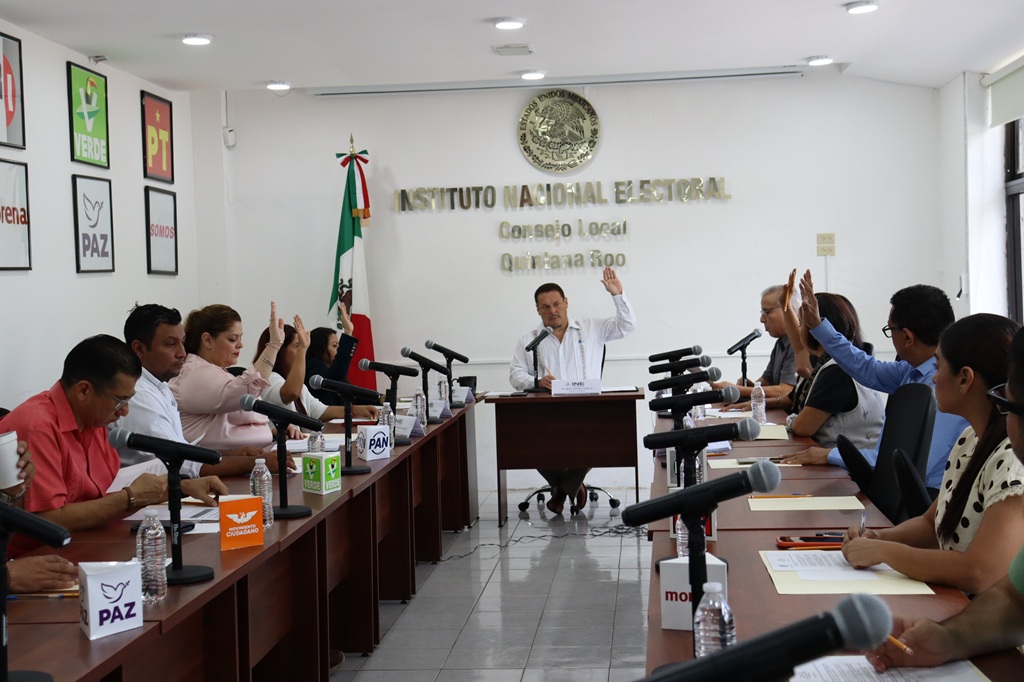 personas sentadas en un salón color blanco votando a mano alzada