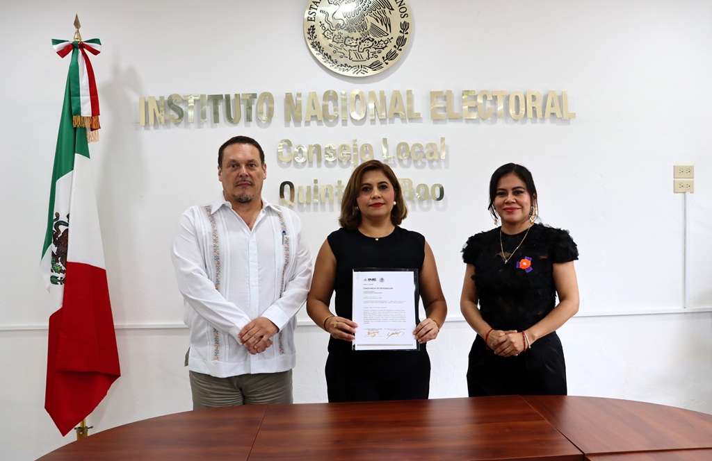 tres personas de pie, una mujer enmedio sostiene un diploma