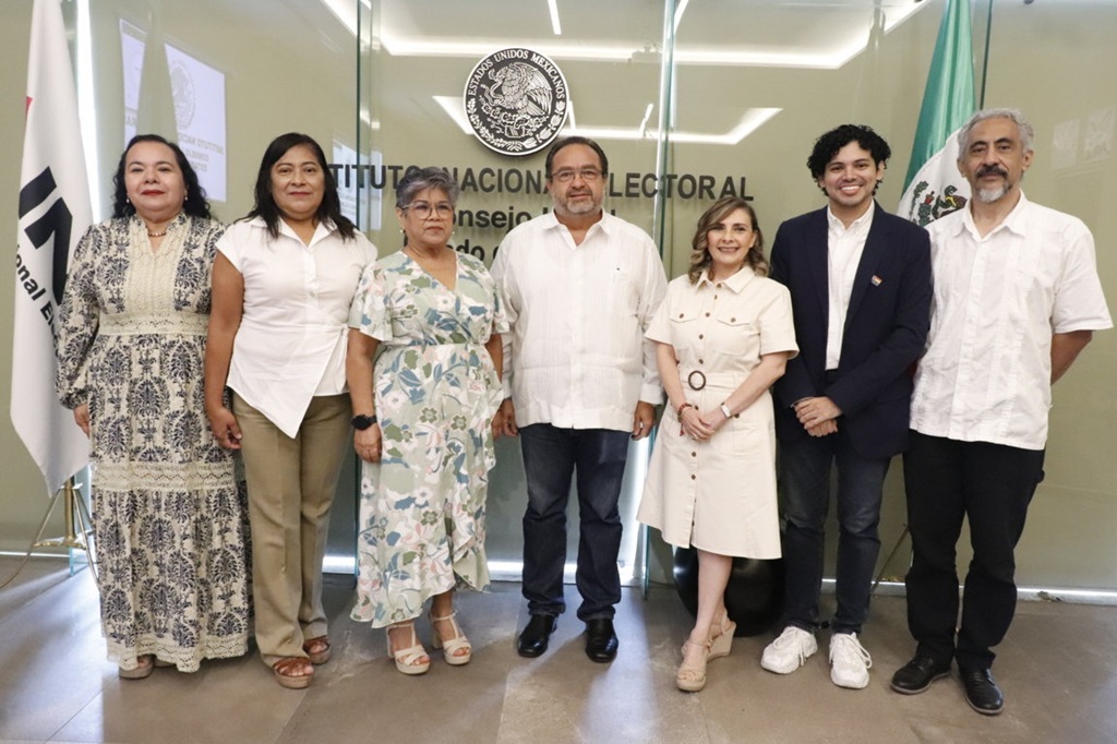 cuatro mujeres y tres hombres de pie frente a una pared de cristal con el escudo nacional
