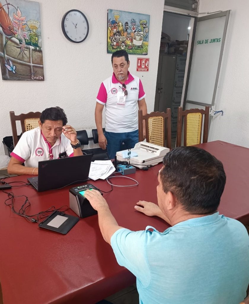 Tres hombres uno coloca mano en maquina digital , otro frente a una computadora y uno más de pie