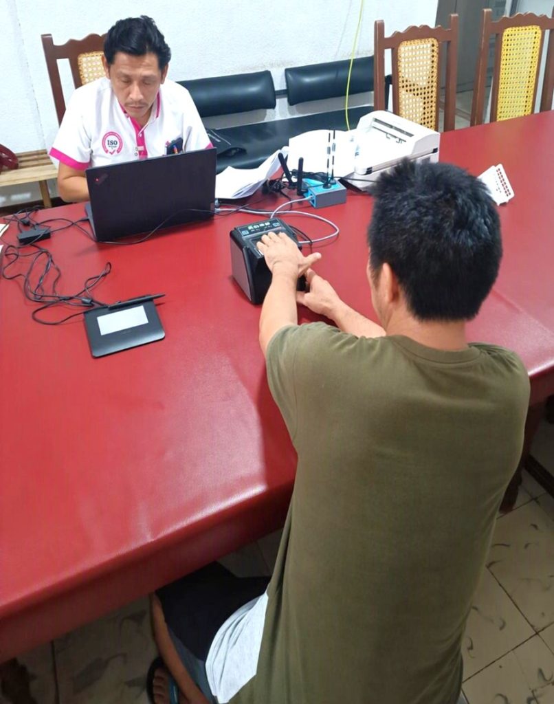 dos hombres sentados, uno frente a computadora mientras otro coloca huellas digitales en maquina