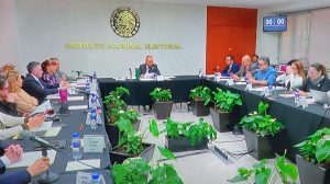 Personas en reunión en una sala de juntas con un escudo de la República Mexicana al fondo